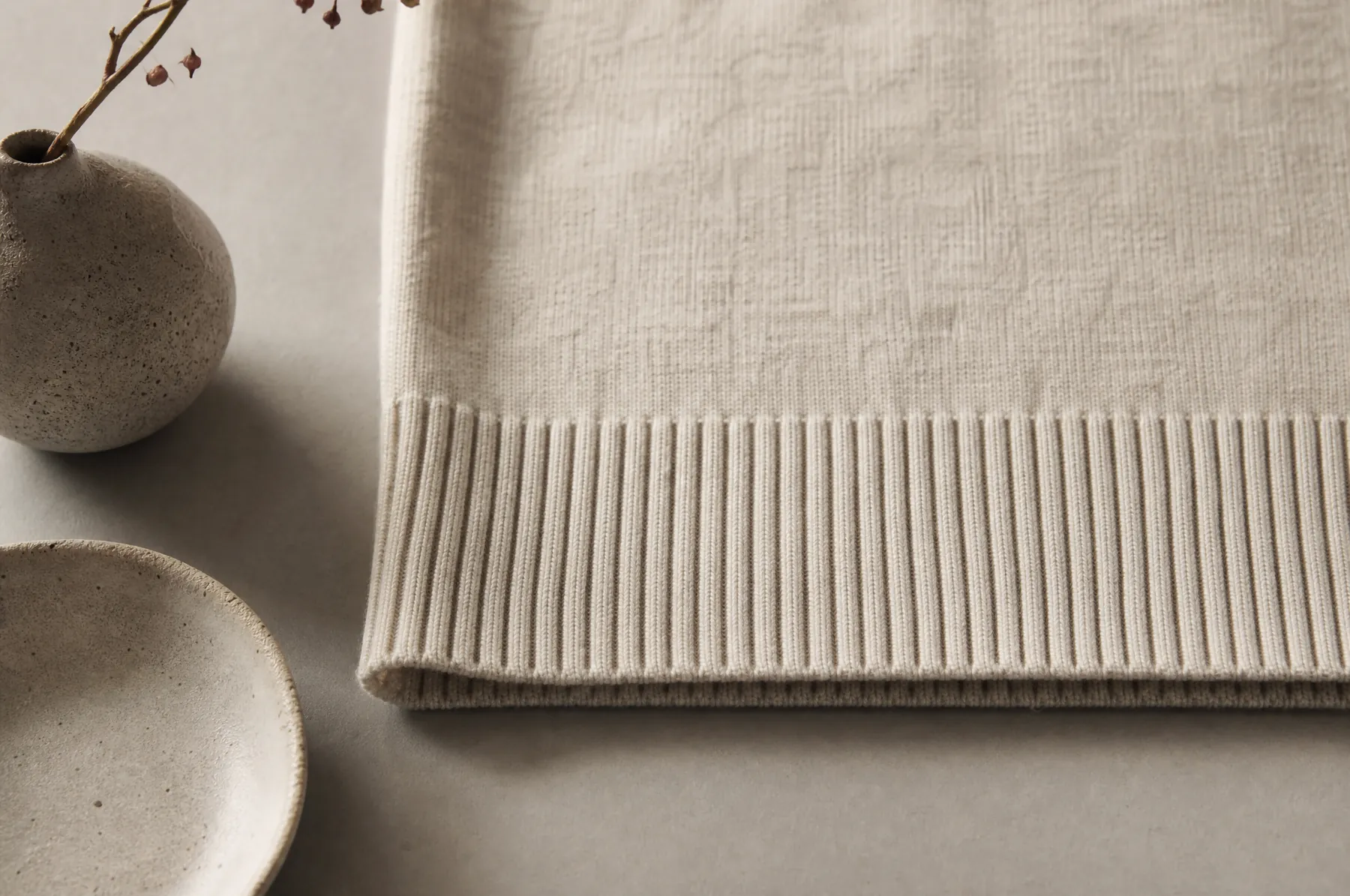 The ribbed hem of a fine oat-coloured knit laid flat on a pale paper ground.