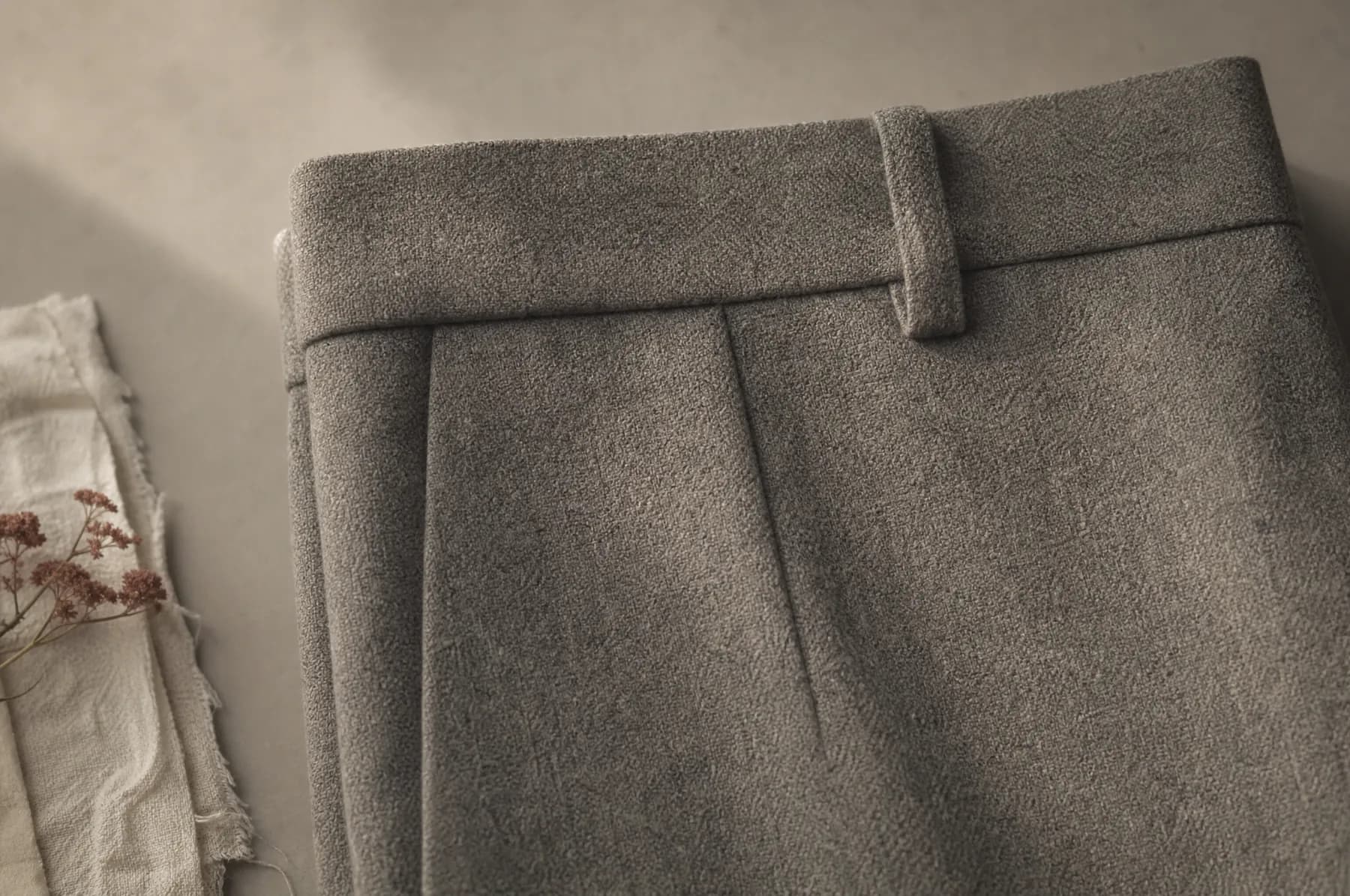 Wool trousers folded flat on a pale paper ground with the waistband and one belt loop in frame.